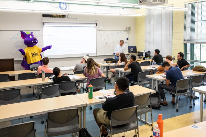 The ACC Riverbat mascot presenting to a classroom of students seated at tables.