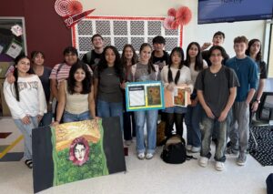 A group of students posing together in a classroom displaying their student art projects.