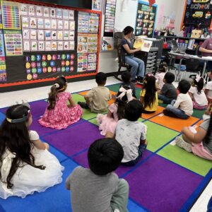 Students sitting on a classroom rug listening to an instructor read a picture book.