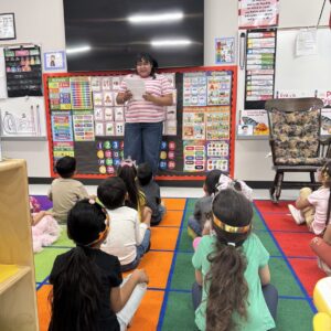 An instructor reading aloud to a group of elementary students sitting on a colorful classroom rug.
