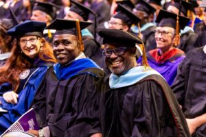 Faculty members wearing academic regalia with doctoral caps and blue hoods smiling together at a graduation ceremony.